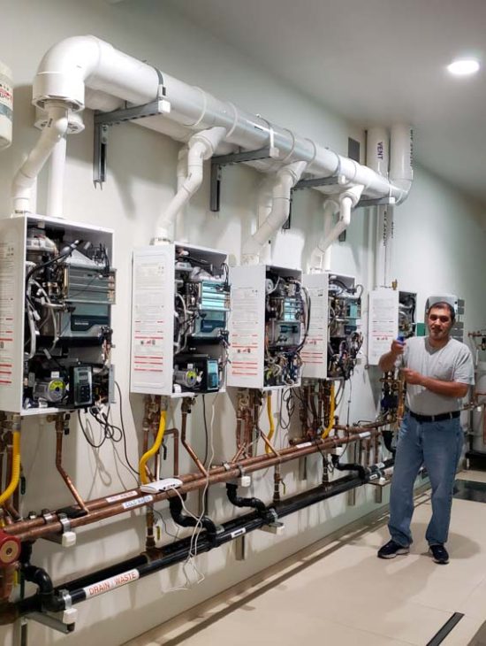 A plumbing instructor standing in a workshop with multiple tankless water heaters mounted for educational purposes.