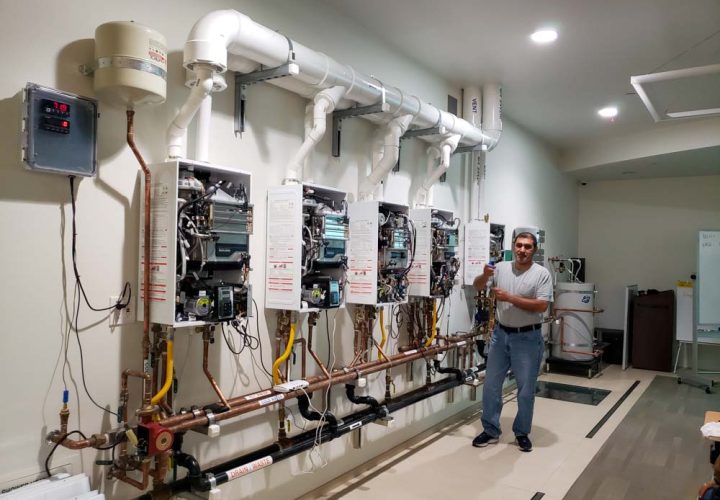 A plumbing instructor standing in a workshop with multiple tankless water heaters mounted for educational purposes.