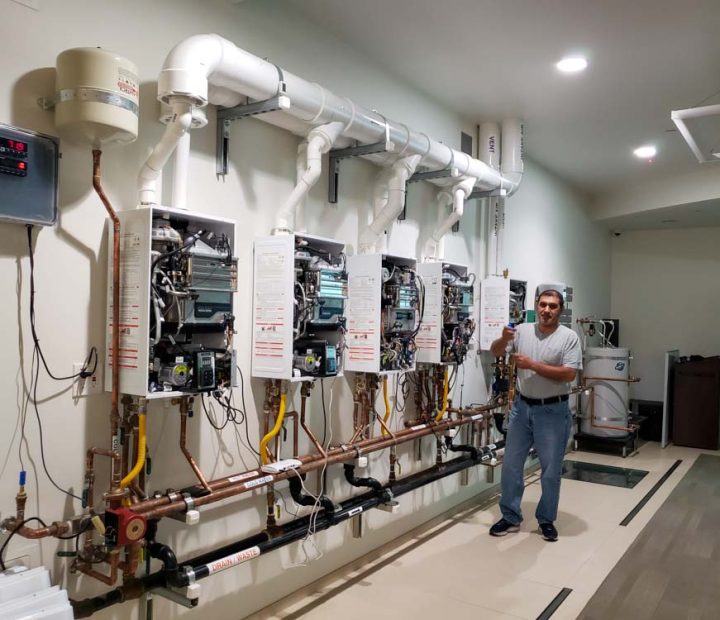 A plumbing instructor standing in a workshop with multiple tankless water heaters mounted for educational purposes.