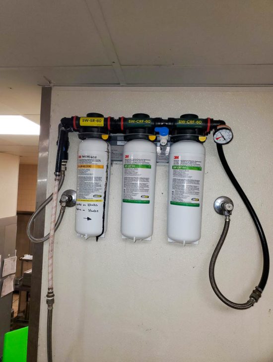 Industrial water filtration system mounted on a wall with three large white canisters, equipped with pressure gauges and metal piping.