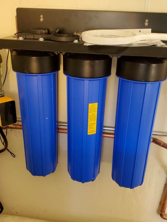 Three large blue water purification tanks installed in a utility room, connected to a complex array of pipes and gauges.