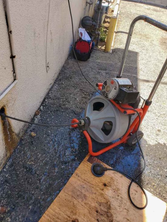 Portable electric drain cleaning machine in operation outside, showing cables and water supply connected, ready for heavy-duty use.