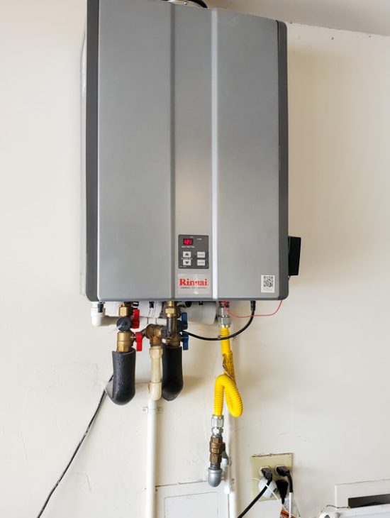 Rinnai tankless water heater installed on a white wall in a garage, with gas and water connections neatly organized.