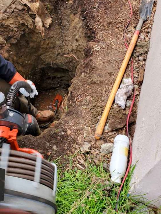Deep excavation revealing underground plumbing, with a plumber working amidst dirt and debris.