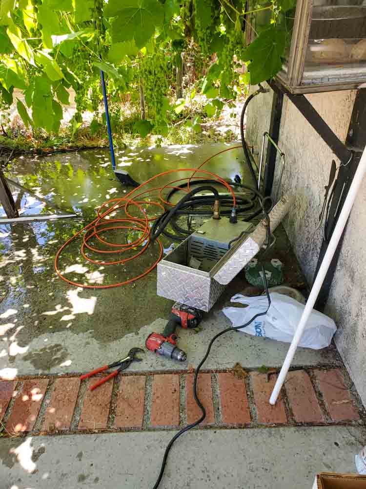 An outdoor plumbing setup featuring various tools and a flooded area under grapevines, indicating active work.
