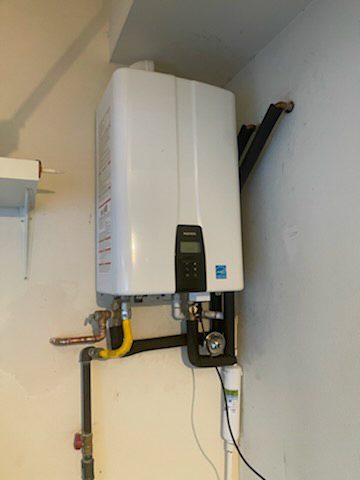 Compact white tankless water heater mounted on a light gray wall, connected to plumbing fixtures.