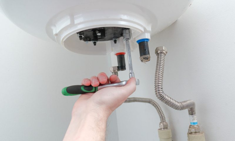 A plumber's hand using a wrench to adjust connections on a white water heater, focusing on precision and safety.