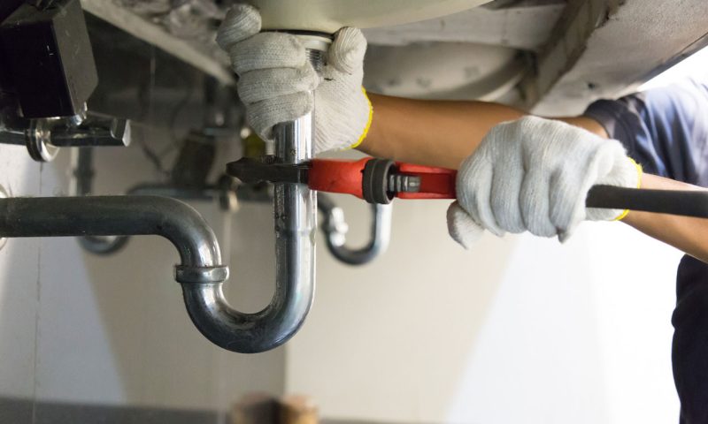 A plumber working to fix a sink's plumbing, using wrenches on the pipes beneath the sink